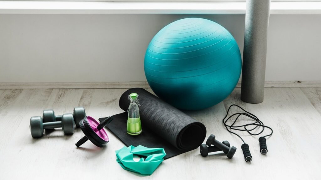 "Small gym equipment including adjustable dumbbells, resistance bands, kettlebell, yoga mat, jump rope, and foam roller arranged in a compact home gym."